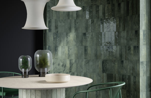 A modern dining area with glossy green rectangular tiled walls, a round light-colored table, two green glass vases, green metal chairs, and two large white pendant lights above.