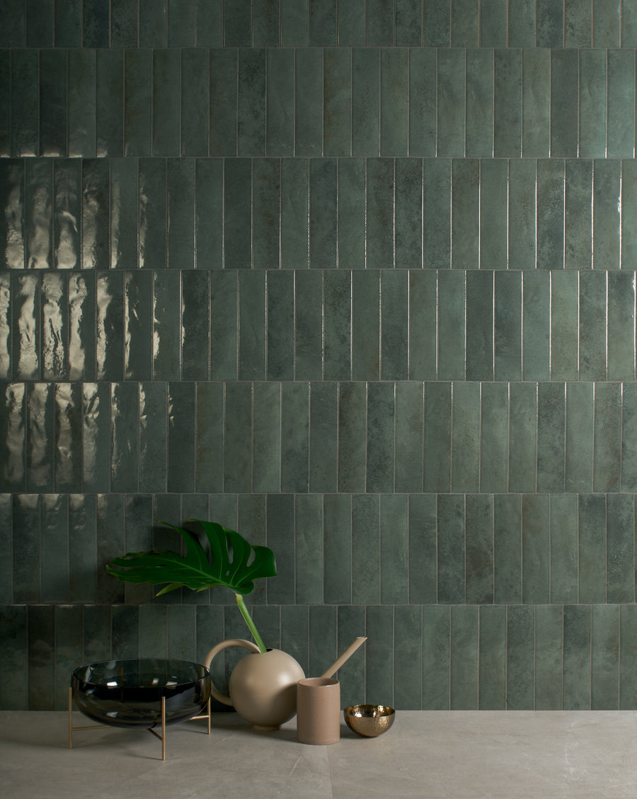A modern setting with glossy green rectangular tiles arranged vertically on a wall, and a beige table holding a green leaf in a vase, a black bowl, a beige cup, and a small gold bowl.