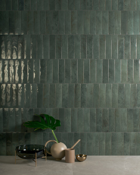 A modern setting with glossy green rectangular tiles arranged vertically on a wall, and a beige table holding a green leaf in a vase, a black bowl, a beige cup, and a small gold bowl.