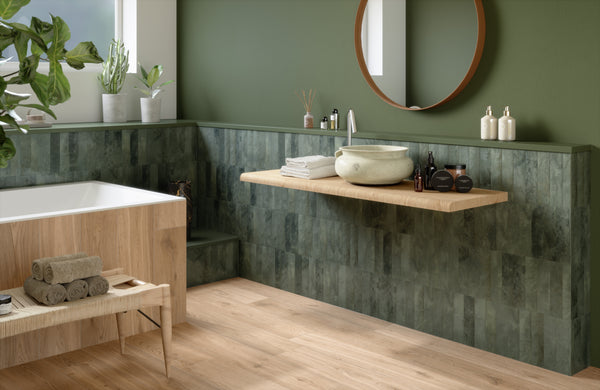 Modern bathroom with green walls, wood and stone accents, a round mirror, vessel sink, rolled towels, and various toiletries on a floating wooden vanity, and a wooden bathtub. Natural light and plants add a fresh feel.