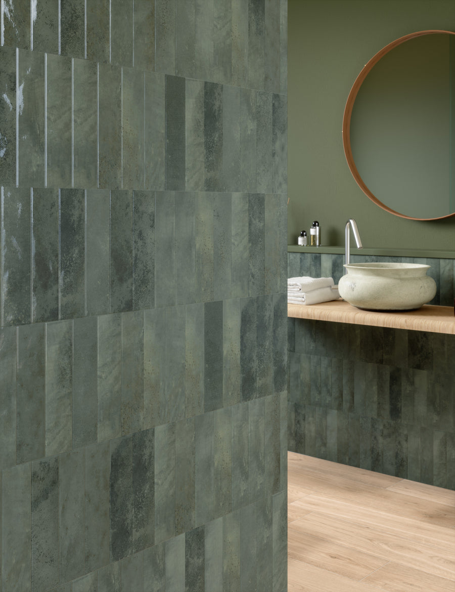 Modern bathroom with green rectangular tiles on the wall, a round mirror above a light wood countertop, a stone vessel sink, toiletries, and neatly folded white towels. Light wood flooring is visible.