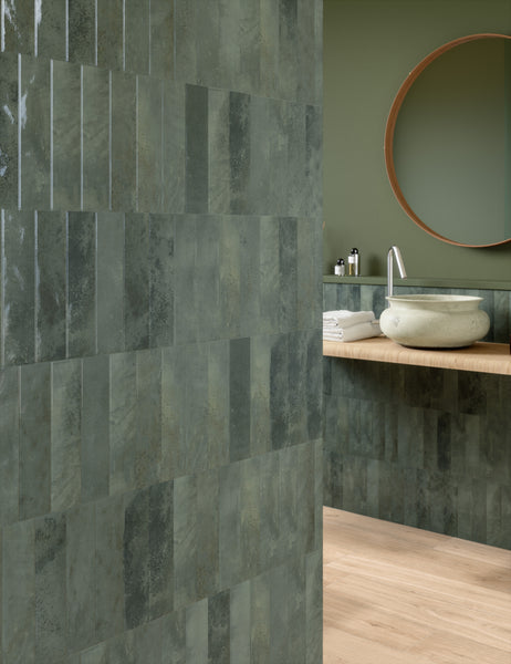 Modern bathroom with green rectangular tiles on the wall, a round mirror above a light wood countertop, a stone vessel sink, toiletries, and neatly folded white towels. Light wood flooring is visible.