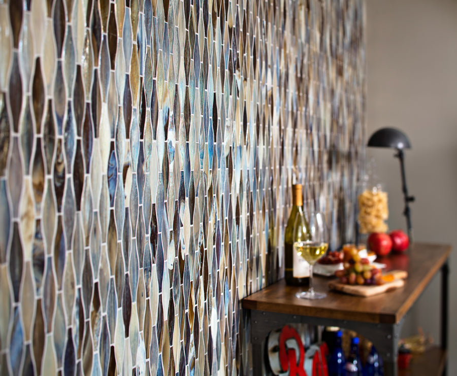 A close-up of a wall with multicolored geometric tile patterns beside a wooden bar cart holding wine, fruit, snacks, a wine glass, and a black desk lamp.