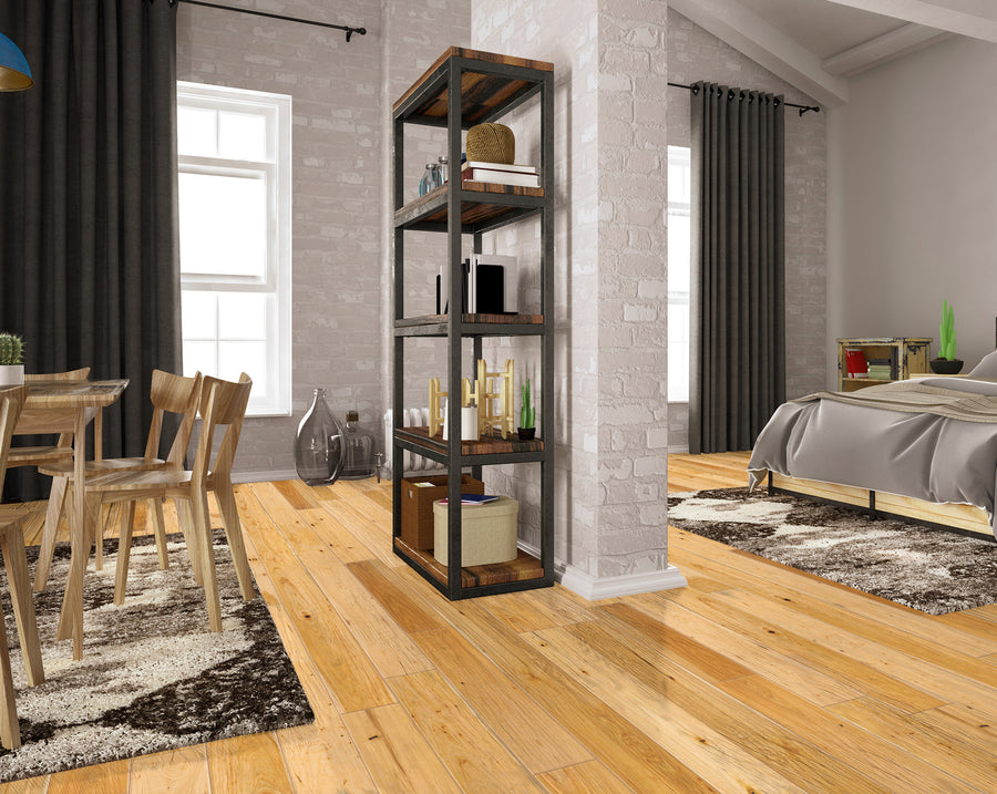 A modern bedroom and dining area separated by a brick pillar with a wooden shelf. The room has light wood floors, neutral rugs, large windows with dark curtains, a small dining table, and a neatly made bed.