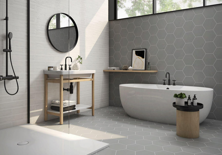 Modern bathroom with a freestanding white bathtub, hexagonal gray tiles on the wall and floor, a round mirror above a sink with an open wooden vanity, and a small round table holding toiletries and a potted plant.