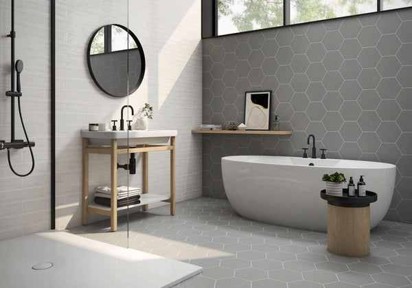Modern bathroom with a freestanding white bathtub, hexagonal gray tiles on the wall and floor, a round mirror above a sink with an open wooden vanity, and a small round table holding toiletries and a potted plant.