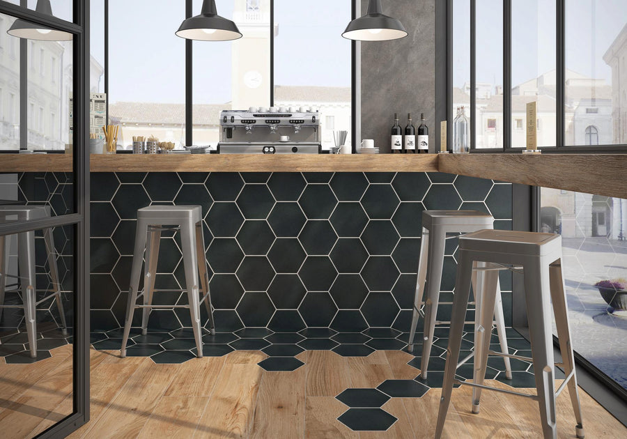 A modern café with tall stools by a wooden counter, hexagonal black tiles on the bar front and floor, large windows, hanging lights, and bottles of wine on the counter.