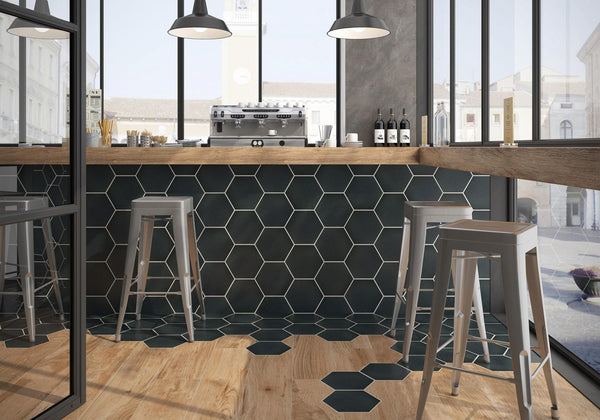 A modern café with tall stools by a wooden counter, hexagonal black tiles on the bar front and floor, large windows, hanging lights, and bottles of wine on the counter.