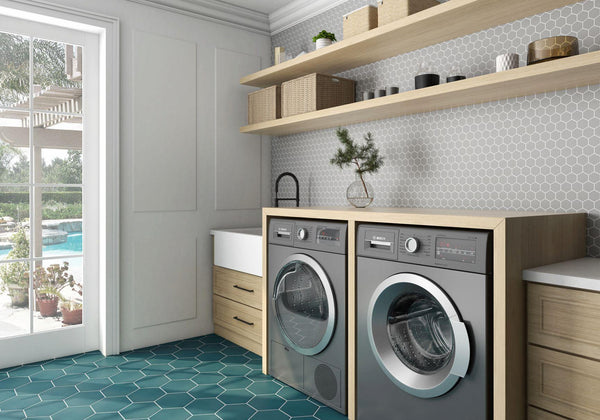 Modern laundry room with hexagon tile floor, two front-loading machines, wooden cabinets, open shelves with baskets and decor, a farmhouse sink, and large window showing a pool and patio outside.
