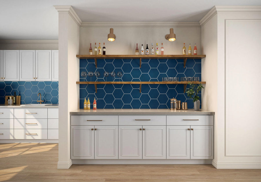 A home bar area with white cabinets, a blue hexagonal tile backsplash, two wooden shelves holding bottles and glasses, and two wall-mounted lights above. The kitchen is partially visible in the background.