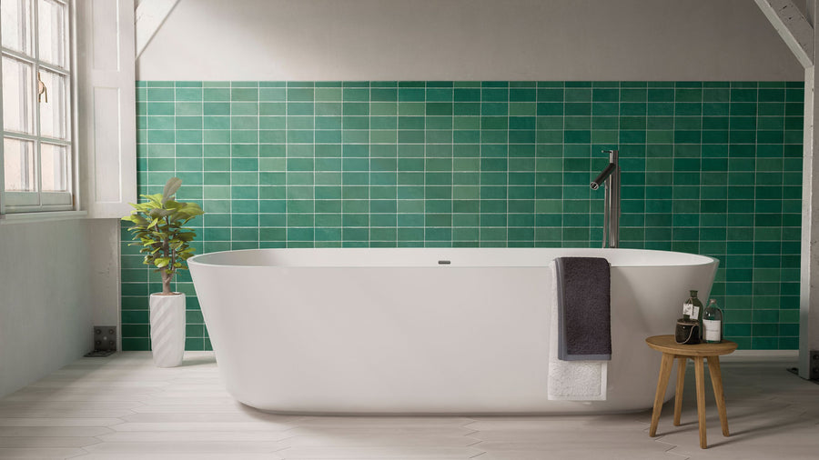 A modern bathroom with a white freestanding bathtub, green tiled wall, a plant in a tall vase, a small wooden stool holding bottles and glasses, and a towel draped over the tub’s edge. Natural light streams in from a window.