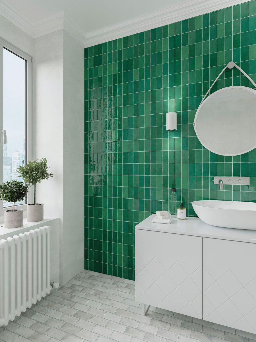 Modern bathroom with a green tiled wall, white vanity with a vessel sink, round mirror, wall sconce, large window, and potted plants on a radiator. The floor is covered with light gray tiles.