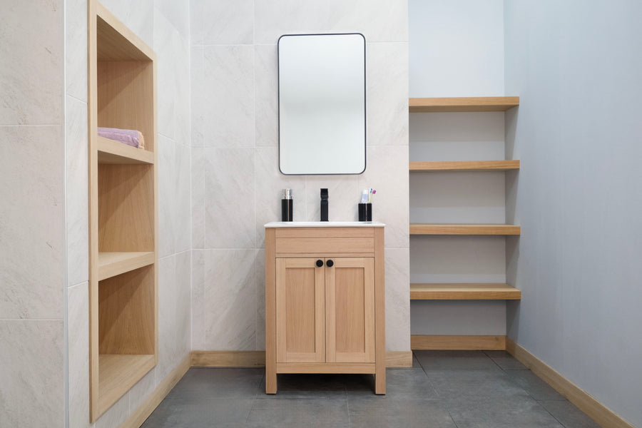 Minimalist bathroom featuring the Aria 24" Vanity with light wood shelves, a rectangular mirror above, toiletries on the counter, and empty wall shelves to the right. Its transitional style blends tiled and painted light walls for a seamless look.