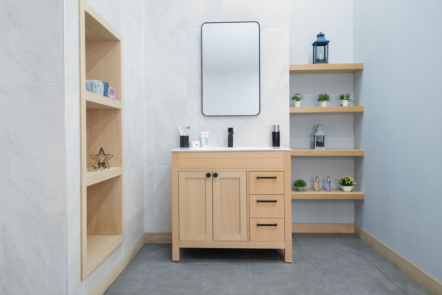 A modern bathroom with an Aria 36" Vanity in light wood, open shelves, a rectangular mirror above the sink, toiletries on the counter, and plants and decor on built-in shelves. The space has light walls and gray tile floors for a transitional look.