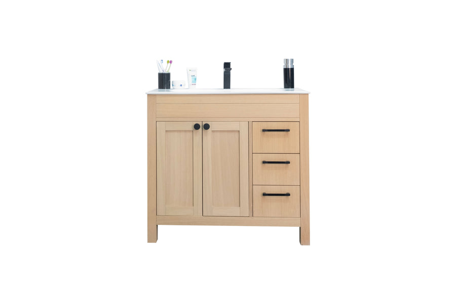 A light wood bathroom vanity with two cabinet doors, three drawers with black handles, and a white countertop holding toiletries, a toothbrush holder, and a soap dispenser.