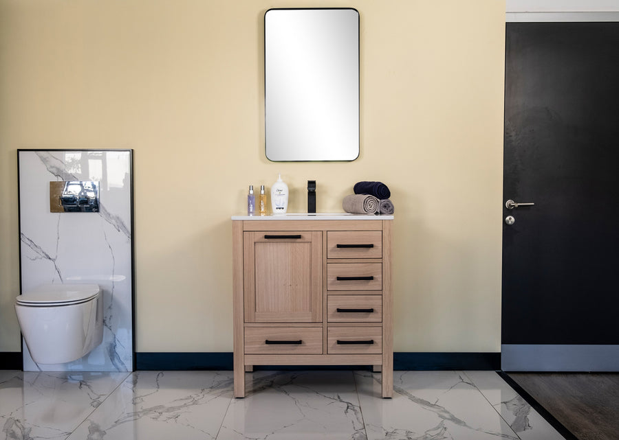 A modern bathroom features the Ashley 30" Vanity in natural oak at the center, with toiletries and towels organized, a wall-mounted toilet to the left, a rectangular mirror above, black door on the right, and white marble tile flooring.