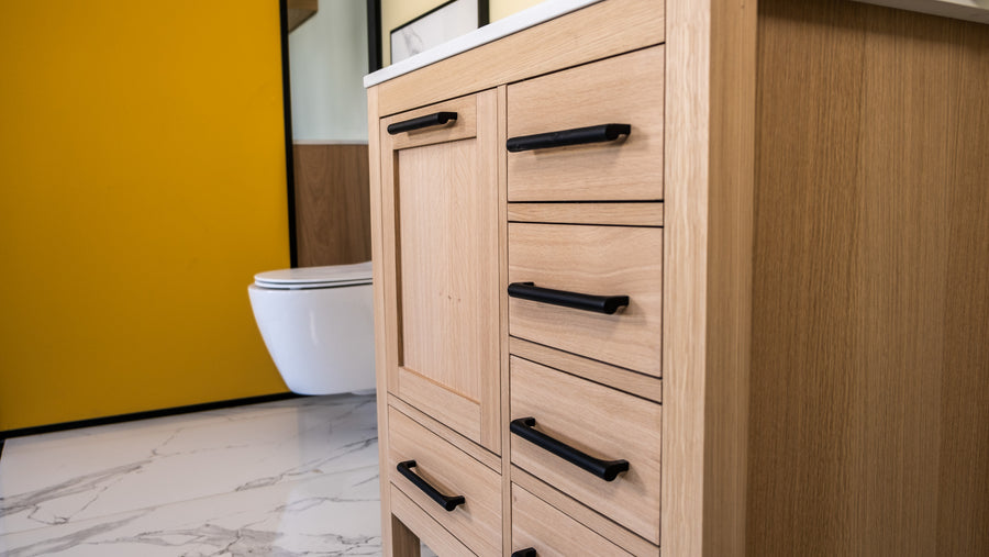 Light wood bathroom vanity with black handles on drawers and cabinet, white countertop, and a modern white toilet in the background. The floor has white marble tiles, and theres a yellow wall panel.
