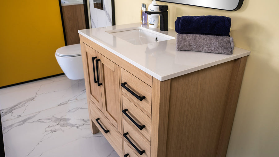 A modern bathroom vanity with a white countertop, a built-in sink, wooden cabinets with black handles, and neatly folded navy and gray towels on top. Marble-patterned floor tiles and a wall-mounted toilet are visible.