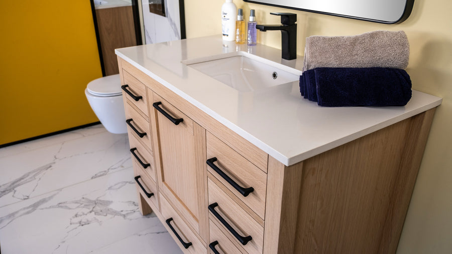 A modern bathroom vanity with a white countertop, black faucet, and wooden drawers. Two folded towels, one gray and one navy, are stacked on the counter. The floor is white with gray veining, and a toilet is visible nearby.