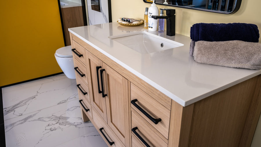 Modern bathroom with a light wood vanity, white countertop and sink, black handles, folded navy and beige towels, toiletries, and a wall-mounted toilet. Marble tile floor and a yellow wall are also visible.