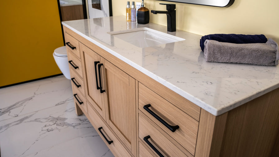Light wood bathroom vanity with a white marble countertop, black hardware, under-mount sink, and black faucet. Towels and toiletries are placed on the counter. Yellow wall and toilet are partially visible.