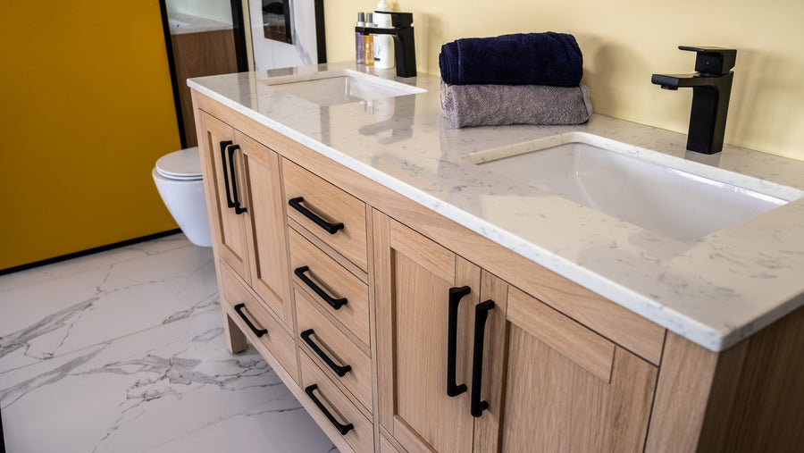 Modern bathroom with a light wood vanity, white marble countertop, black fixtures, double sinks, folded towels, and a toilet in the background. The floor and walls have a clean, minimalist design.