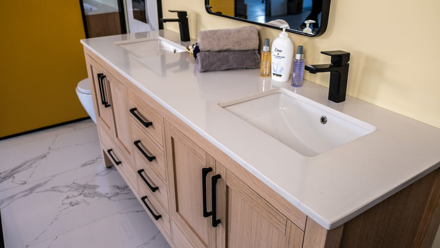 Double bathroom vanity with light wood cabinets, black handles, two sinks, black faucets, a soap dispenser, Dove lotion, toiletries, and folded towels on a white countertop, set against a yellow wall and marble floor.