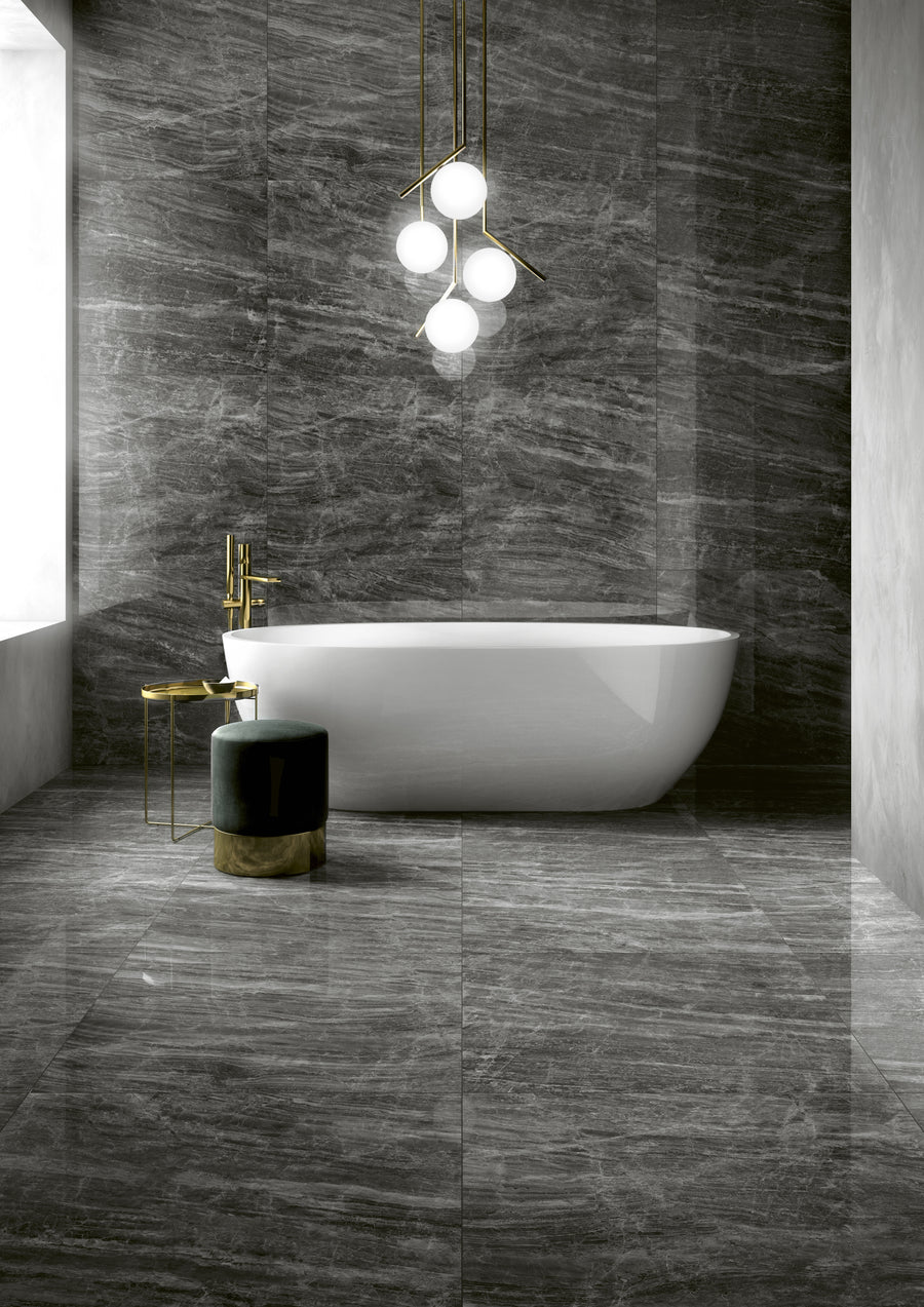 A modern bathroom with dark gray marble floors and walls, a white freestanding bathtub, a gold faucet, a green velvet stool with a gold base, and a contemporary chandelier with globe lights.