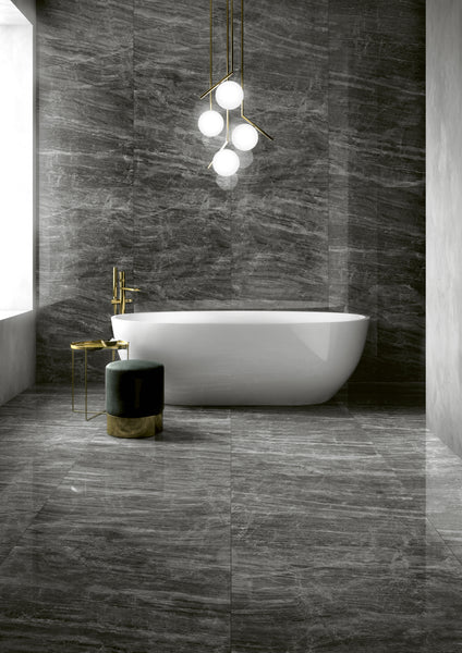 A modern bathroom with dark gray marble floors and walls, a white freestanding bathtub, a gold faucet, a green velvet stool with a gold base, and a contemporary chandelier with globe lights.