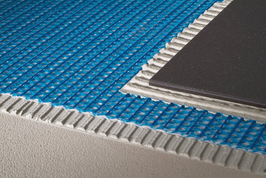 A close-up of a floor installation shows a Blanke Permat 2' x 3.2" Sheet used as blue uncoupling underlayment over adhesive, with a black tile partially set atop the membrane and adhesive layers.