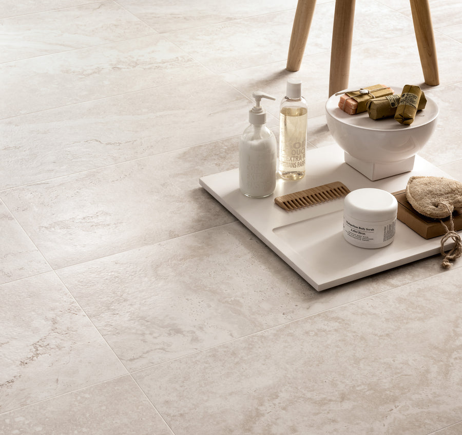 A tray holding lotion, soap bars, a comb, a jar, and a sponge rests on light beige stone-tile flooring next to wooden stool legs. The setting appears clean and spa-like.