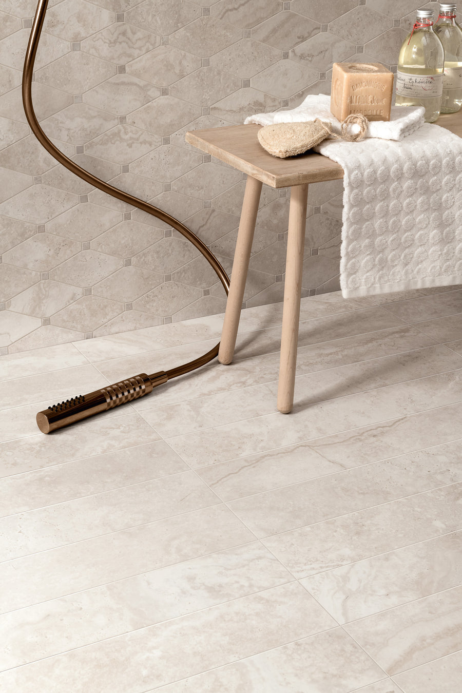 A modern bathroom scene with beige tiled walls and floors, a wooden stool holding folded towels, soap, a body brush, sponges, and glass bottles of water. A bronze handheld showerhead lies on the floor.