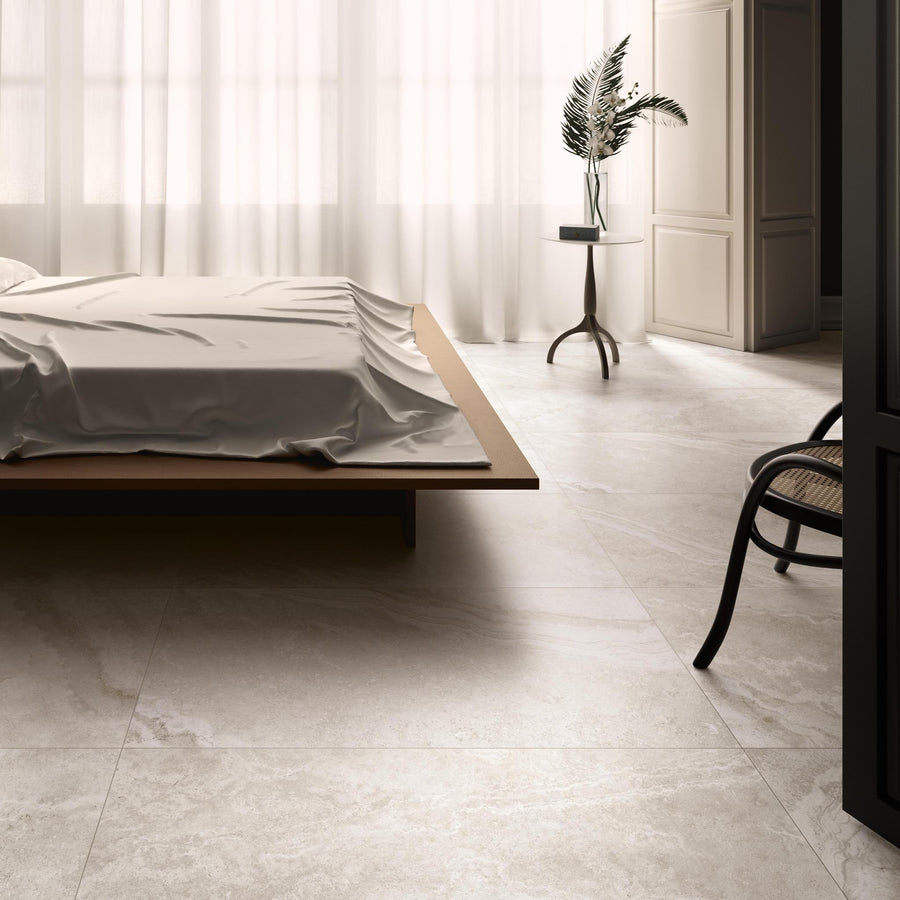 Minimalist bedroom with large marble tile floor, a low wooden bed with rumpled sheets, sheer white curtains, a round table holding a vase with leafy branches, and a dark wooden chair in the corner. Soft natural light fills the room.