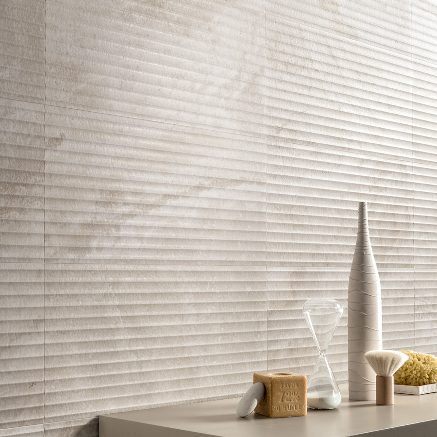 Minimalist bathroom scene with textured beige tiled walls. On a light countertop, there is a decorative white bottle, an hourglass, a bar of soap, a white brush, and a natural sponge.
