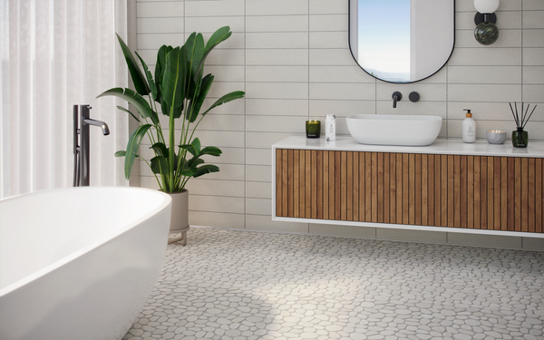 Modern bathroom with a white freestanding tub, a vanity with wood paneling, a round mirror, a potted plant, and pebble-style tile flooring. Natural light streams in through sheer curtains.