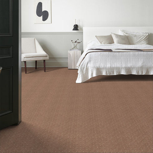 A minimalist bedroom with a white bed, neutral bedding, light beige chair, a vase on a small table, modern wall art, a black wall lamp, and light brown carpet flooring. The walls are white with wainscoting.