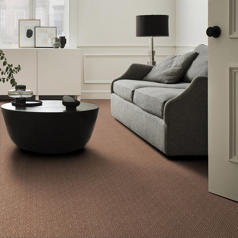 A modern living room with a grey sofa, black round coffee table, white sideboard, lamp, and minimal decor. The floor is covered with a patterned brown carpet. Light streams in from a window out of frame.