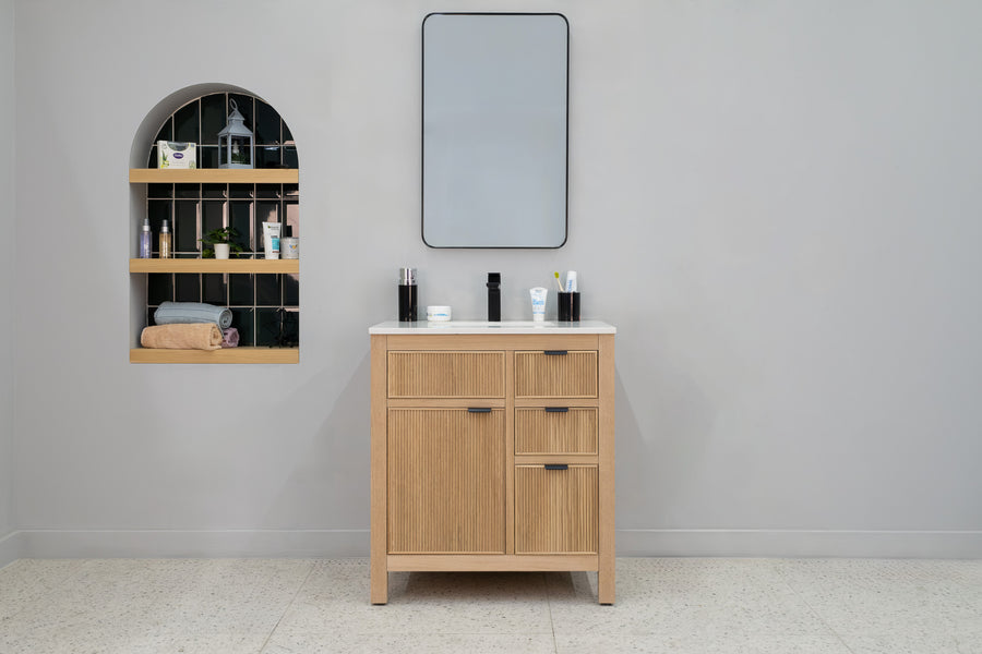 A modern bathroom features the Catalina 30" Vanity with a white countertop, a rectangular mirror, and open wall shelves displaying toiletries, plants, and rolled towels. Light gray walls complete the transitional look.