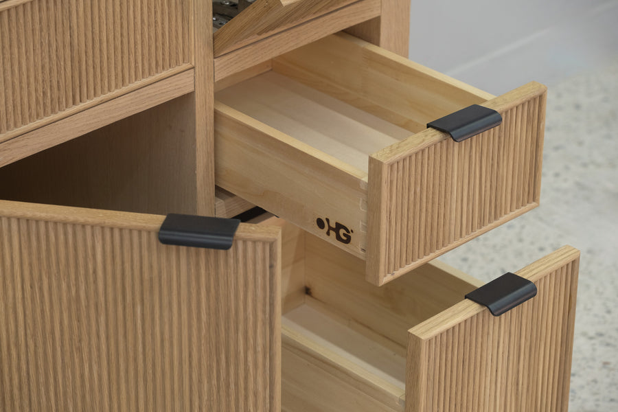 A close-up view of a wooden cabinet with several drawers partially open, featuring light wood grain and black rectangular handles, placed on a speckled floor.