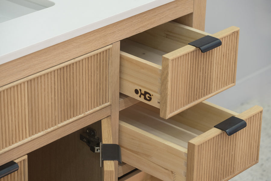 A close-up of a wooden bathroom vanity with two open drawers, black handles, and a vertical groove design. The cabinet doors below are also slightly open, revealing the interior.