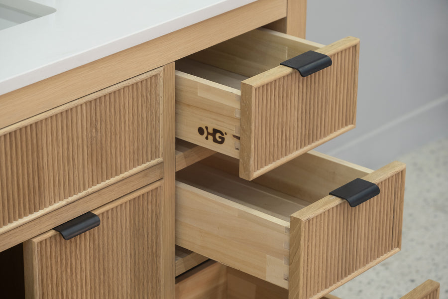 A close-up of a wooden cabinet with two drawers partially open, showing light wood interiors and black handles. The cabinet has a grooved texture and a white countertop above.