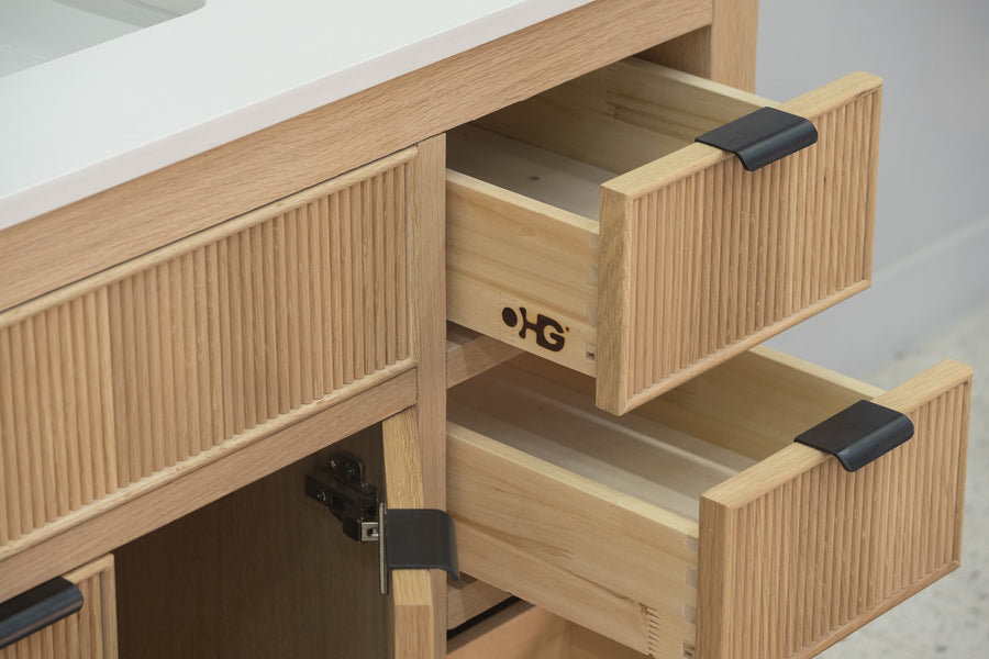 Close-up of a wooden cabinet with light wood grain finish, showing two open drawers and a partially open door. The drawers have black handles and inner branding that reads “OHG.”.