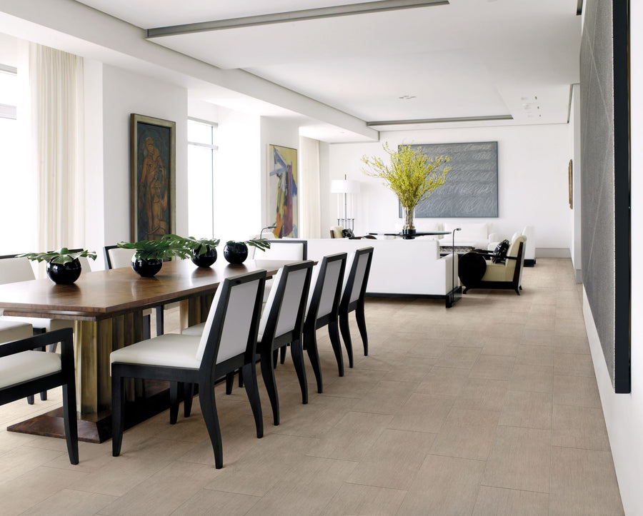 Modern open-plan dining and living room with a wooden dining table, black and white chairs, potted plants, artwork on the walls, and a white sofa with a vase of yellow flowers on a table. Light wood floors throughout.