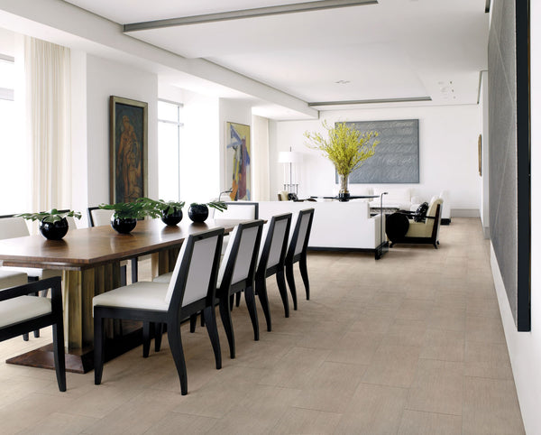 Modern open-plan dining and living room with a wooden dining table, black and white chairs, potted plants, artwork on the walls, and a white sofa with a vase of yellow flowers on a table. Light wood floors throughout.