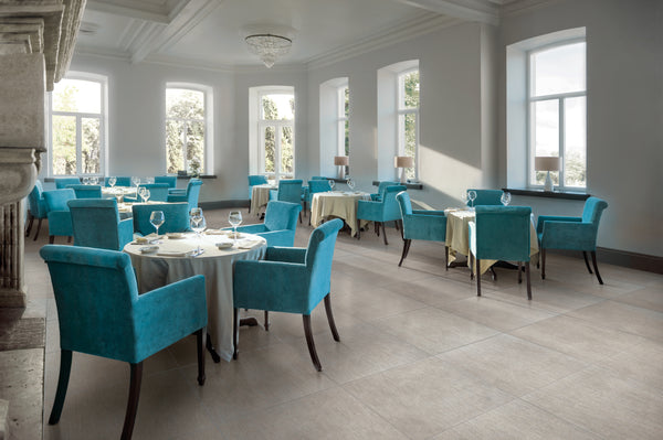 Bright, elegant restaurant dining room with large windows, turquoise upholstered chairs, round tables set with white cloths, glassware, and cutlery. Natural light fills the space, creating an inviting atmosphere.