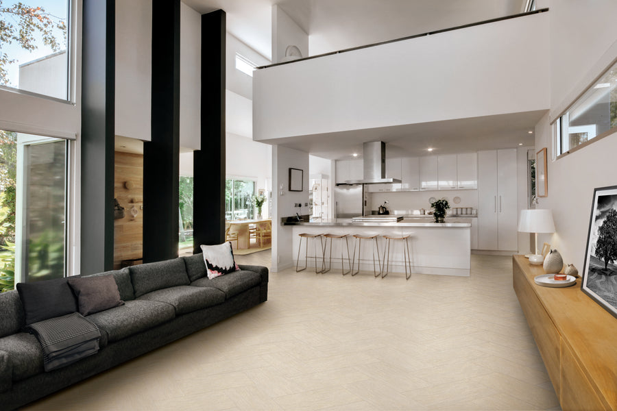 Modern open-plan living space with high ceilings, a dark gray sofa, large windows, a white kitchen with bar stools, and minimalist decor, including framed art and plants. Bright and airy atmosphere.