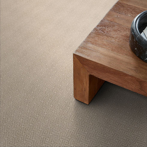 Close-up of a beige carpet with a subtle geometric diamond pattern next to the corner of a wooden coffee table and a round black decorative bowl.