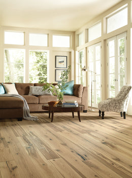A cozy living room with large windows features a tan sectional sofa, blue pillows, a patterned armchair, and a round wooden coffee table with a plant. Sunlight streams onto Montauk 7