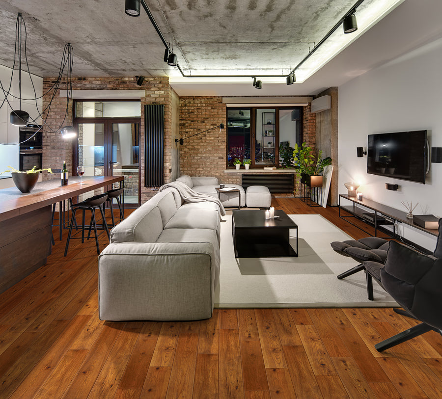 Modern open-plan living room and kitchen with exposed brick walls, wooden flooring, gray sectional sofa, black chairs, wall-mounted TV, and industrial-style lighting. A large window brings in natural light.