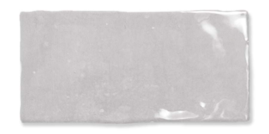 Rectangular, glossy light gray ceramic tile with slightly uneven edges and a smooth, reflective surface. The shine creates subtle highlights and variations in tone across the tile.